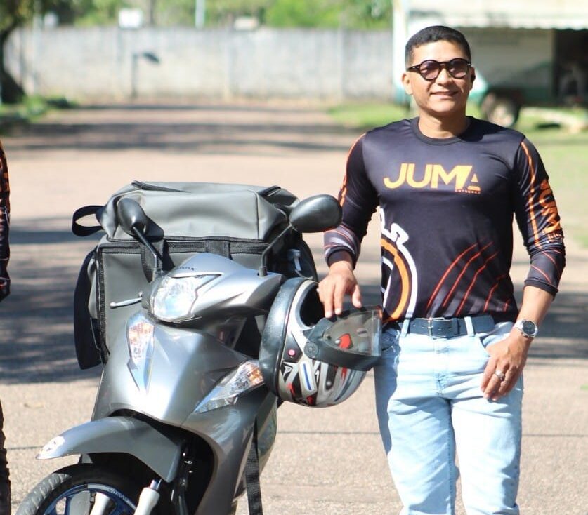 Homem, Robson Pantoja, entregador da Juma Entregas, sorrindo ao lado de uma moto.