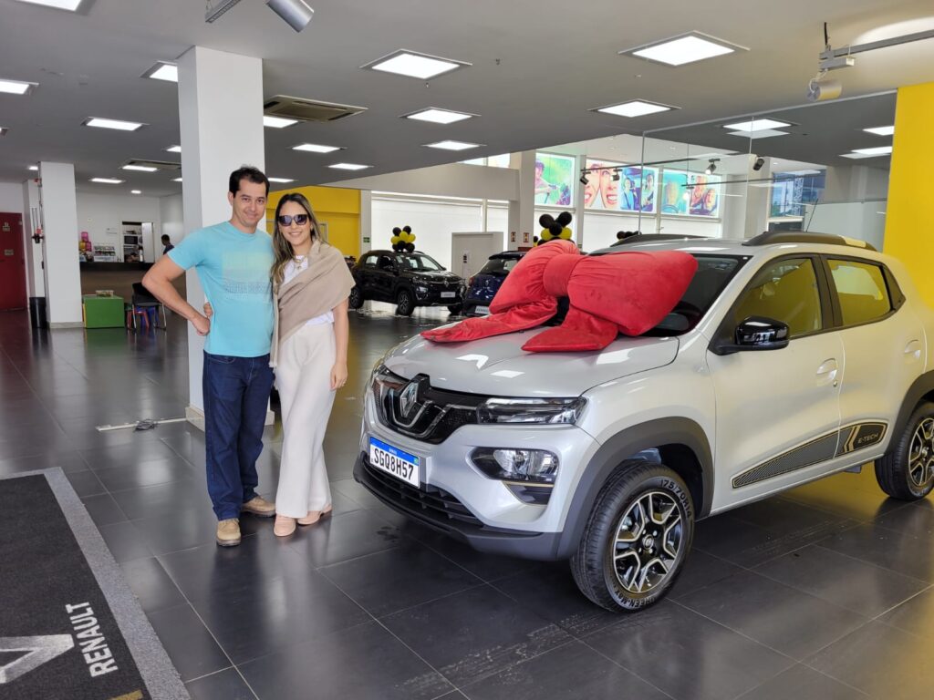 Casal posando ao lado de um carro Renault Kwid E-Tech branco com laço vermelho no capô dentro de uma concessionária.