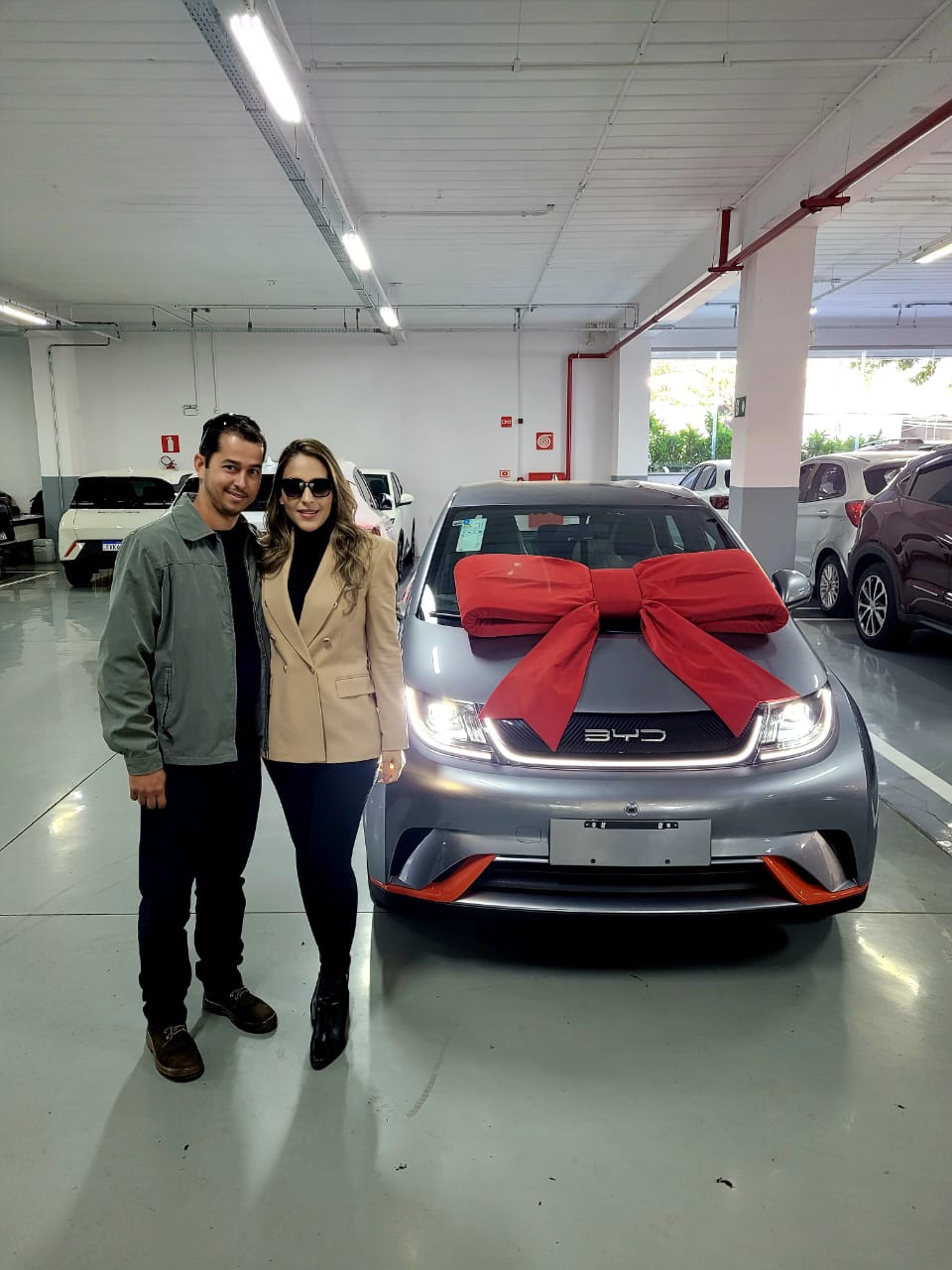 Casal posando ao lado de um carro BYD prata com detalhes laranja e laço vermelho no capô, em estacionamento coberto de concessionária.