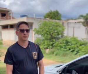 Homem de camiseta preta com óculos escuros em frente a uma área residencial com vegetação e muro ao fundo.