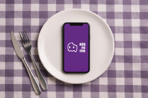 Mesa quadriculada com celular mostrando a tela do aiqfome em cima de um prato, e talheres ao lado.