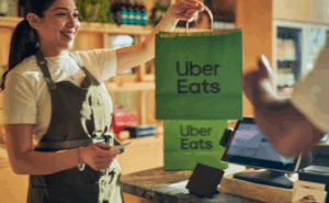 Funcionária de restaurante sorrindo enquanto entrega sacola verde do Uber Eats a um cliente no balcão.