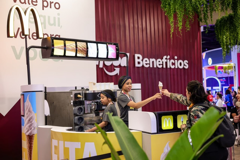 Loja do McDonald's, com logo do iFood Benefícios ao fundo, e atendente entregando um sorte de casquinha a uma cliente.