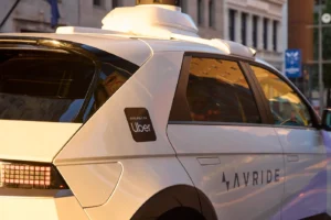 Carro autônomo branco com logotipo da Uber e da Avride estacionado em área urbana ao entardecer.