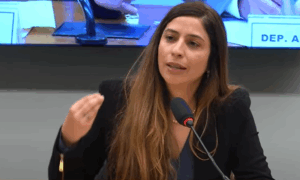 Mulher fala ao microfone durante sessão em ambiente formal, com gesto de mão levantada e expressão séria.