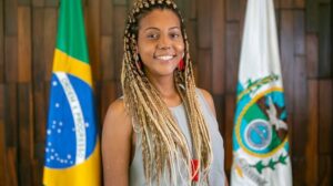 Mulher sorridente com tranças loiras posando em frente às bandeiras do Brasil e do estado do Rio de Janeiro.