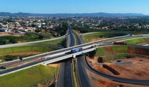 Fotografia aérea de um entroncamento rodoviário em Montes Claros, Minas Gerais, com viadutos e pistas largas cercadas por áreas verdes e de terra, e a cidade ao fundo.