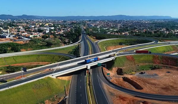 Fotografia aérea de um entroncamento rodoviário em Montes Claros, Minas Gerais, com viadutos e pistas largas cercadas por áreas verdes e de terra, e a cidade ao fundo.