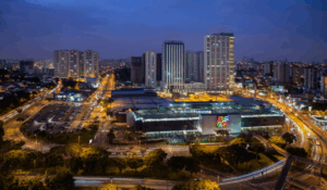 "Vista noturna de um centro urbano iluminado, com prédios altos ao fundo e um grande shopping ao centro, cercado por ruas movimentadas com tráfego de carros."