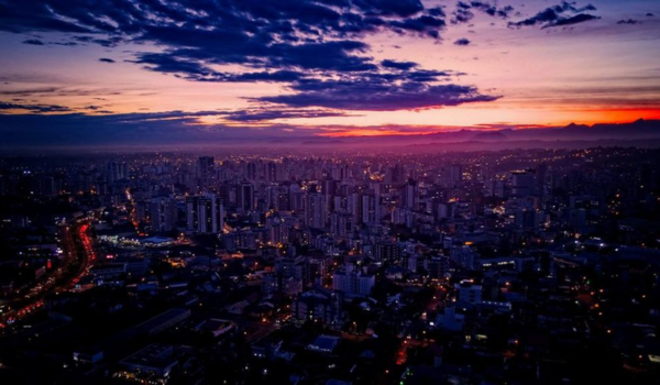 Vista aérea de uma grande cidade ao anoitecer, com céu em tons de roxo, azul e laranja, prédios iluminados e ruas com tráfego de carros visíveis pelas luzes vermelhas e brancas.