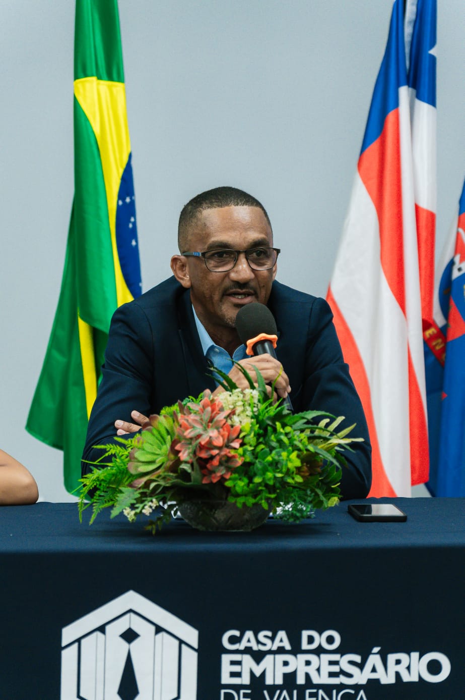 Homem falando ao microfone durante um evento institucional, com bandeiras do Brasil, Bahia e outra ao fundo.