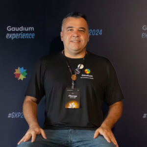 Homem sorrindo, usando camiseta preta com o logotipo da Gaudium e crachá identificado como “Pitanga”, posando em frente a um painel do evento Gaudium Experience 2024.