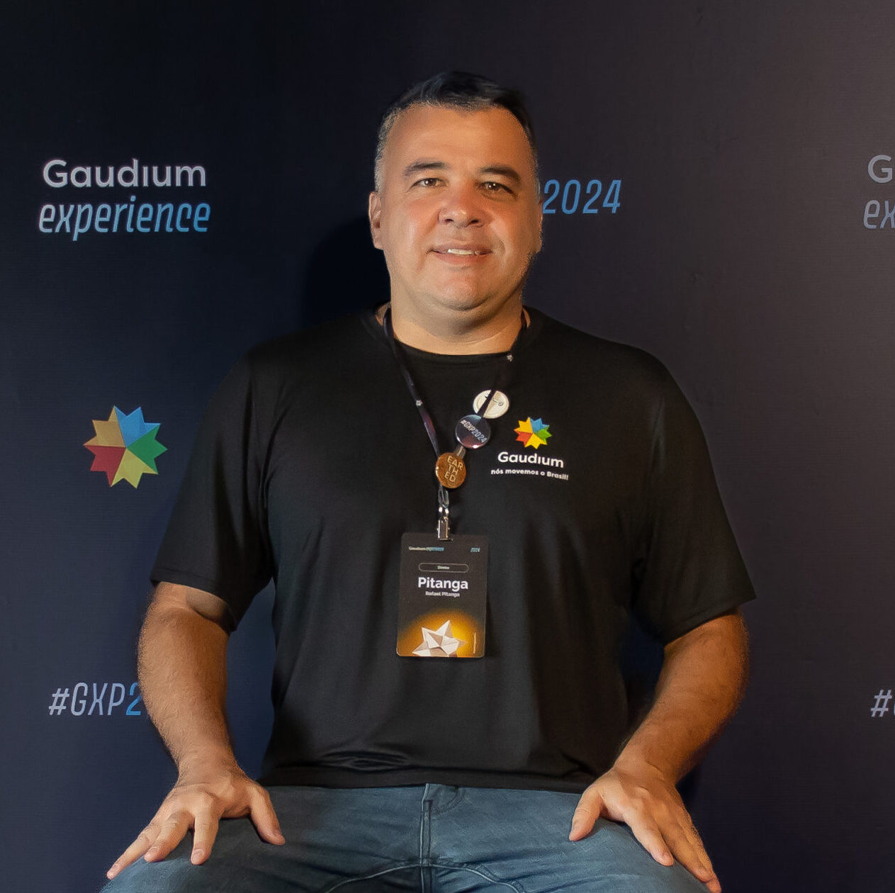 Homem sorrindo, usando camiseta preta com o logotipo da Gaudium e crachá identificado como “Pitanga”, posando em frente a um painel do evento Gaudium Experience 2024.