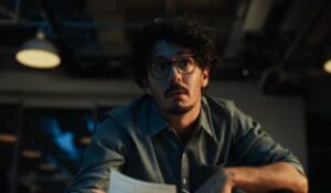 Homem de óculos e cabelo encaracolado olha surpreso enquanto segura um papel em um ambiente de escritório com iluminação artificial.