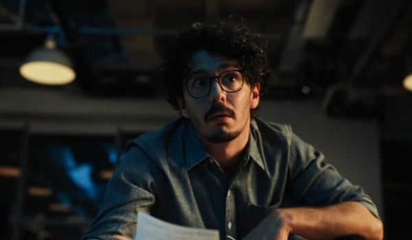 Homem de óculos e cabelo encaracolado olha surpreso enquanto segura um papel em um ambiente de escritório com iluminação artificial.