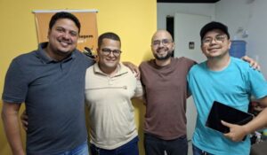 Quatro homens posam sorridentes em frente a uma parede amarela. Eles estão lado a lado, com os braços nos ombros uns dos outros, em um clima de camaradagem.