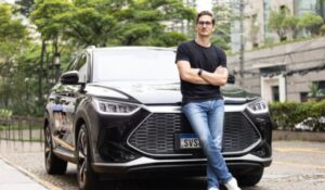 Homem de camiseta preta e calça jeans encostado em um carro preto moderno, em uma rua arborizada com prédios ao fundo.