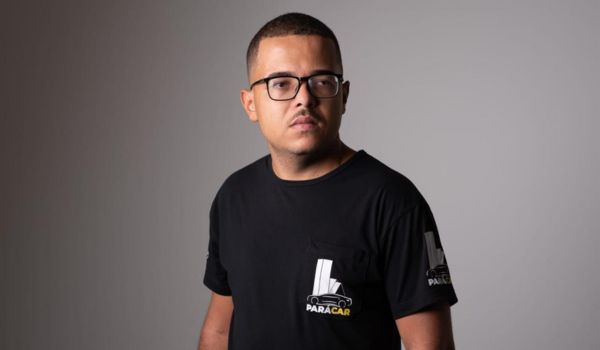Homem de cabelos curtos e barba rala, usando óculos e camiseta preta com o logotipo da empresa “PARACAR”. Ele está olhando ligeiramente para o lado direito, com expressão séria, sobre um fundo neutro em tom cinza.