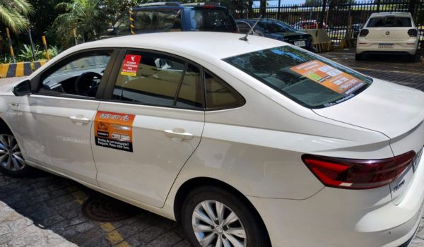 Carro branco estacionado com adesivos publicitários laranja nas portas e no vidro traseiro, em um estacionamento ao ar livre.