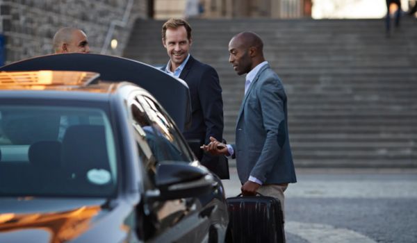 Três homens vestidos com trajes formais estão ao lado de um carro com o porta-malas aberto em uma rua de paralelepípedos. Um deles segura uma mala de viagem enquanto conversa com os outros dois.