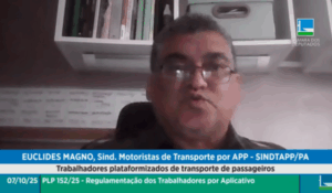 Homem fala por videoconferência durante audiência pública da Câmara dos Deputados, com estante e livros ao fundo.