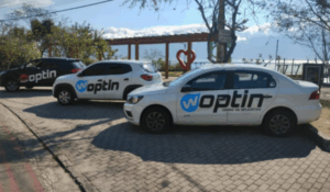 Três carros estacionados em uma rua de paralelepípedos, todos com adesivos grandes da marca Woptin nas laterais, em uma área arborizada com vista para o mar.