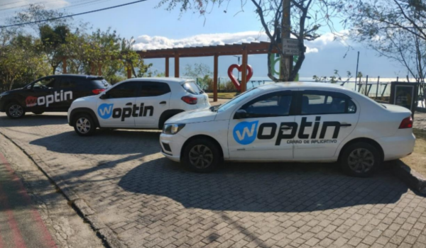 Três carros estacionados em uma rua de paralelepípedos, todos com adesivos grandes da marca Woptin nas laterais, em uma área arborizada com vista para o mar.