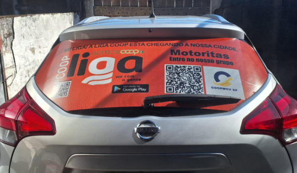 Carro prateado com um adesivo laranja grande no vidro traseiro, divulgando a cooperativa Liga Coop. O adesivo contém o logotipo da Liga Coop, QR codes, o texto “Se liga! A Liga Coop está chegando à nossa cidade” e um convite para motoristas entrarem no grupo.