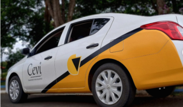 Carro branco com detalhes em amarelo e preto estacionado em uma via arborizada. O veículo exibe a marca “Cevi” e o slogan “Transporte que revoluciona” em sua lateral.