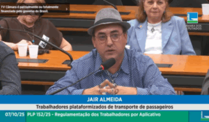 Homem de chapéu e óculos fala ao microfone durante audiência pública na Câmara dos Deputados, com outras pessoas ao fundo.