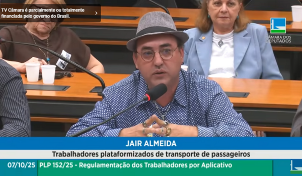 Homem de chapéu e óculos fala ao microfone durante audiência pública na Câmara dos Deputados, com outras pessoas ao fundo.