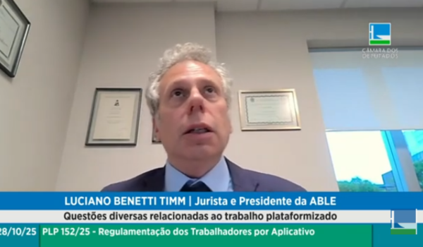 Homem de terno azul fala durante videoconferência de audiência pública sobre regulamentação de trabalhadores por aplicativo, com certificados pendurados na parede ao fundo.
