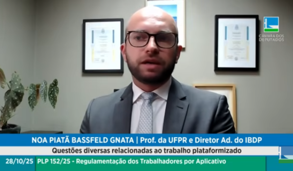Homem de terno e gravata fala em videoconferência durante audiência pública sobre regulamentação de trabalhadores por aplicativo, com certificados emoldurados na parede ao fundo e logotipo da Câmara dos Deputados no canto da imagem.