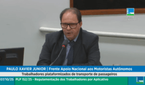 Homem de terno escuro e óculos fala ao microfone durante audiência pública na Câmara dos Deputados. Na legenda, aparece seu nome, “Paulo Xavier Junior”, e sua afiliação à Frente de Apoio Nacional aos Motoristas Autônomos.
