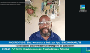 Homem de óculos e barba fala por videoconferência durante audiência pública da Câmara dos Deputados, com medalhas esportivas penduradas ao fundo.
