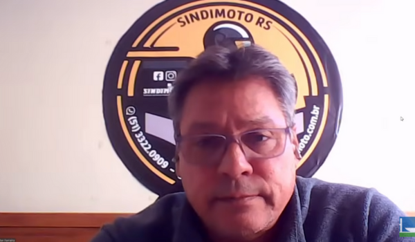 Homem de óculos e expressão séria aparece em videoconferência com o logotipo do Sindimoto RS ao fundo.