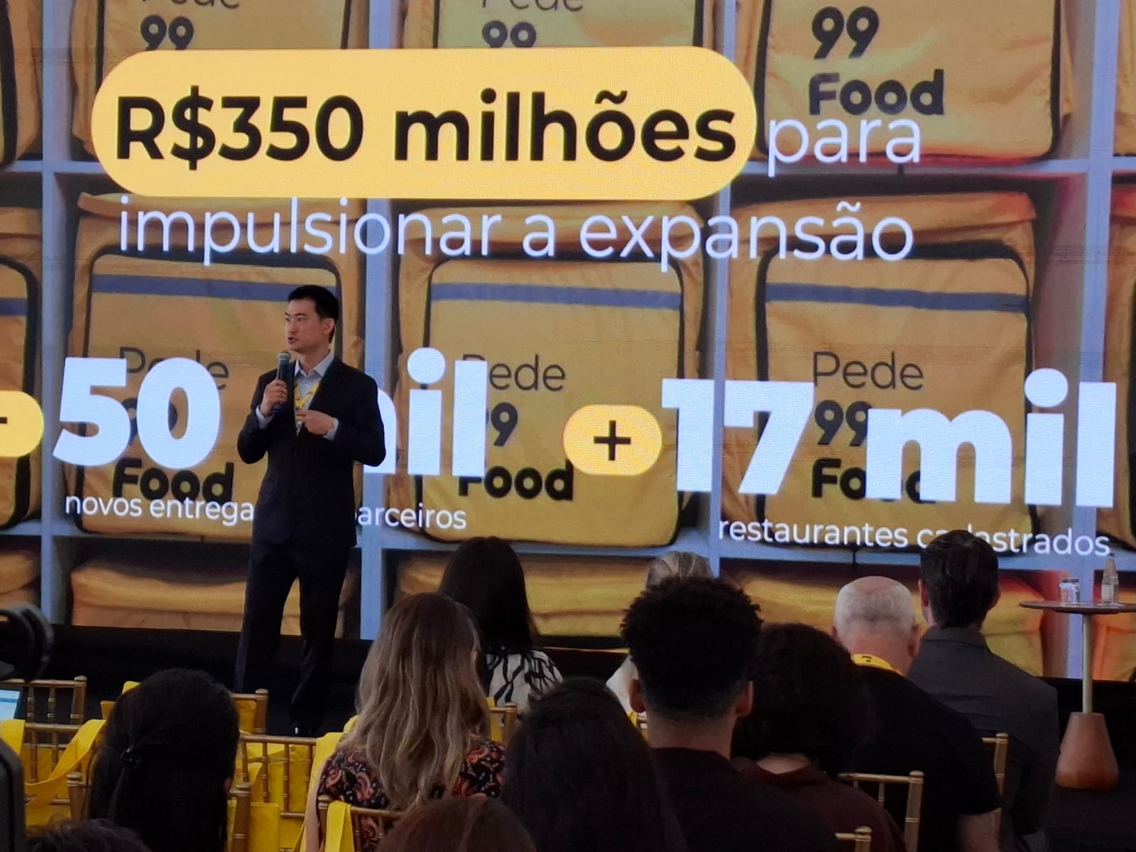 Homem de terno falando ao microfone em um palco diante de um telão com anúncio de investimento da 99Food, diante de uma plateia.