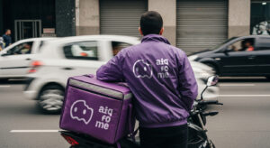 Motorista com jaqueta do aiqfome apoiado em uma bag com a logo da mesma plataforma.