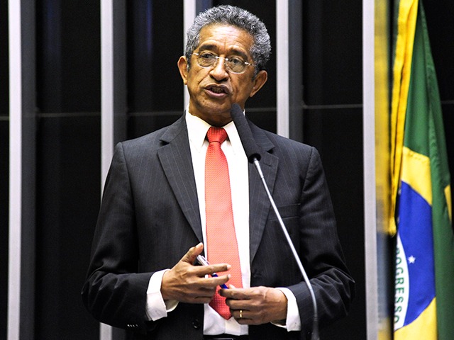 Homem de terno escuro e gravata vermelha fala ao microfone em plenário, com bandeira do Brasil ao fundo.