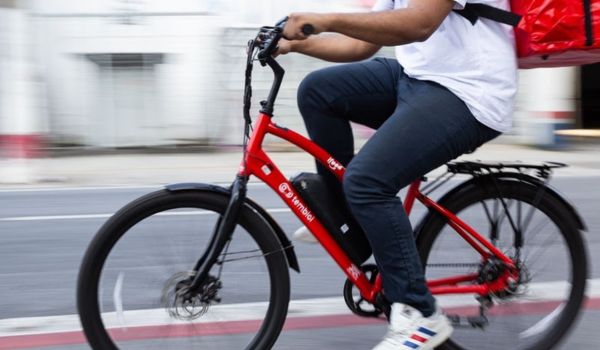 Entregador pedala bicicleta elétrica vermelha do iFood em via urbana. Ele usa mochila de entregas e veste camiseta branca e calça jeans.