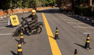 Motociclista com capacete e jaqueta amarela participa de treinamento em pista delimitada por cones, ao lado de placa com o logotipo da 99.