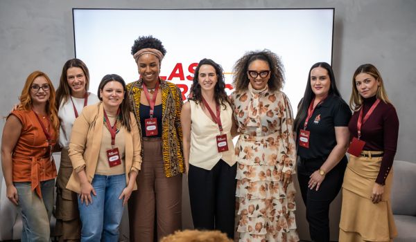 Grupo de nove mulheres posando lado a lado e sorrindo para a câmera em um ambiente corporativo, todas usando crachás de evento.