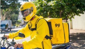 Entregador da empresa 99 em uma motocicleta, usando uniforme e capacete amarelos com o número 99, e carregando uma mochila térmica de entregas da mesma cor.