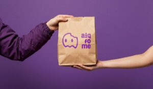 Uma pessoa entrega um saco de papel pardo com o logotipo roxo do aplicativo aiqfome impresso na frente, sobre um fundo roxo.