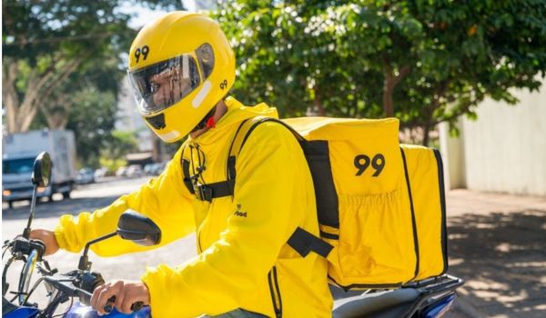 Entregador da empresa 99 em uma motocicleta, usando uniforme e capacete amarelos com o número 99, e carregando uma mochila térmica de entregas da mesma cor.
