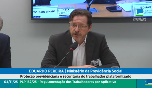 Eduardo Pereira, representante do Ministério da Previdência Social, fala ao microfone durante uma audiência pública na Câmara dos Deputados sobre a proteção previdenciária e securitária do trabalhador plataformizado. Ele usa terno escuro, camisa branca e óculos, sentado à mesa com um crachá visível no paletó.