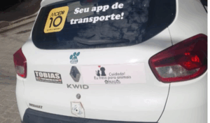 Traseira de um carro branco com adesivos, incluindo “Mobi 10 – Seu app de transporte!” no vidro traseiro e mensagens de campanhas na tampa do porta-malas.