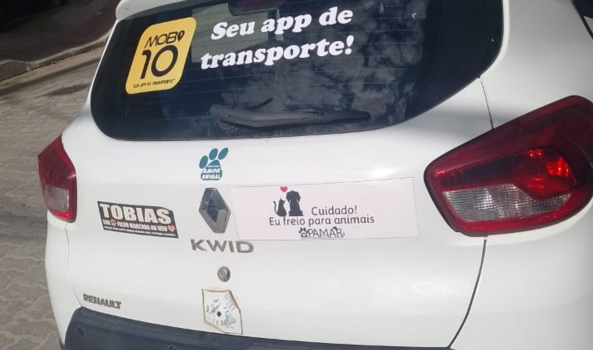 Traseira de um carro branco com adesivos, incluindo “Mobi 10 – Seu app de transporte!” no vidro traseiro e mensagens de campanhas na tampa do porta-malas.