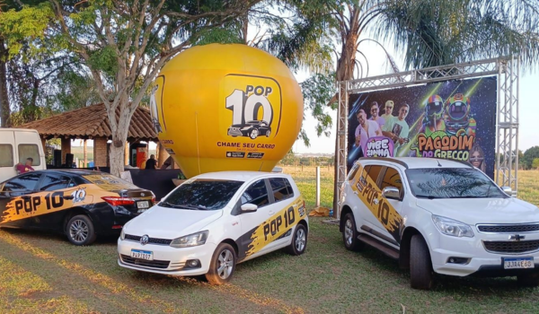 Três carros adesivados com a marca Pop 10 estão estacionados em área aberta com árvores, ao lado de um grande balão amarelo promocional e um painel de evento musical.