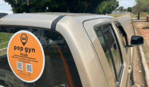 Adesivo circular “pop gyn mob” no vidro traseiro de um carro, com QR codes e texto “Aplicativo goianiense 100% goiano”, em rua arborizada.